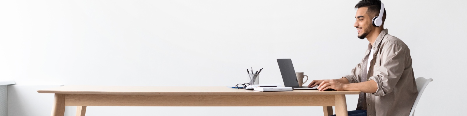 Cheerful Middle Eastern Freelancer Man Working On Pc Online At Home Listening To Music With Wireless Headphones, Making Research For New Project, Sitting At Desk Enjoying Remote Job, Profile Side View